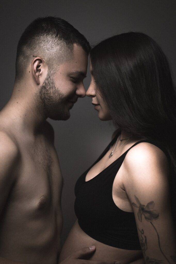 Pareja abrazada, el futuro papá sostiene delicadamente la barriga de su esposa mientras ambos sonríen mirándose con ternura.