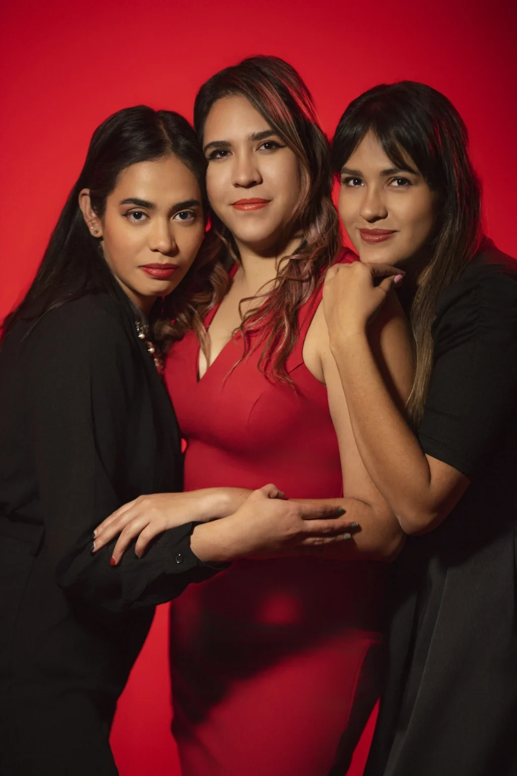 Retrato de estudio de tres mujeres posando juntas frente a un fondo rojo vibrante. La mujer del centro viste un vestido rojo, mientras que las otras dos, a cada lado, llevan atuendos oscuros. Todas tienen cabello oscuro y lápiz labial rojo, mirando directamente a la cámara con expresiones serias pero seguras.