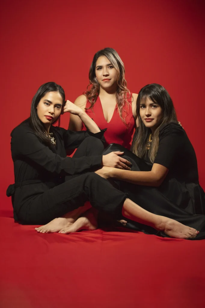 Retrato de estudio de tres mujeres posando juntas frente a un fondo rojo vibrante. La mujer del centro viste un vestido rojo, mientras que las otras dos, a cada lado, llevan atuendos oscuros. Todas tienen cabello oscuro y lápiz labial rojo, mirando directamente a la cámara con expresiones serias pero seguras.