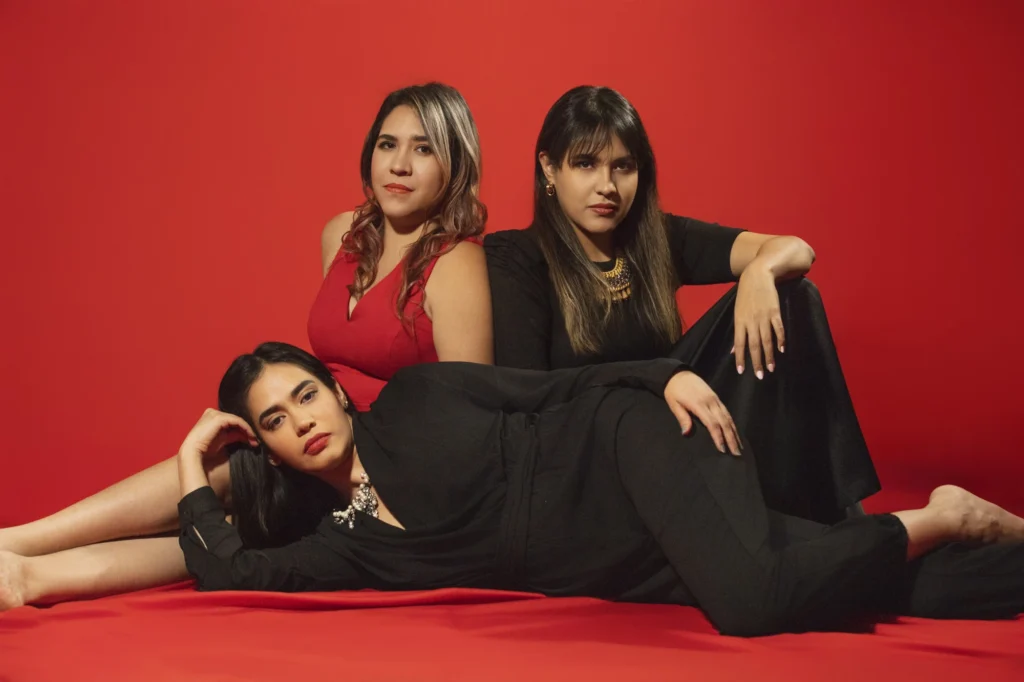 Retrato de estudio de tres mujeres posando juntas frente a un fondo rojo vibrante. La mujer del centro viste un vestido rojo, mientras que las otras dos, a cada lado, llevan atuendos oscuros. Todas tienen cabello oscuro y lápiz labial rojo, mirando directamente a la cámara con expresiones serias pero seguras.